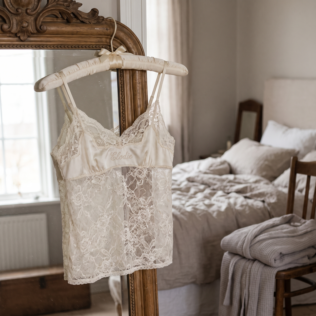 Camisole de dentelle ivoire brodée d'un prénom, suspendue à un cintre rembourré dans une chambre